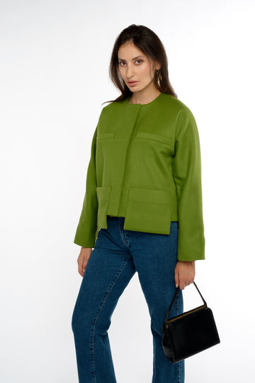 A woman stands in 3/4 view, wearing a modern-cut green lady jacket with drop patch pockets paired with stylish jeans. She holds a vintage black purse, adding a touch of classic elegance to her contemporary outfit. The scene captures her confident stance against a neutral background.