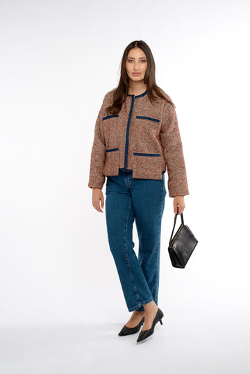 A woman stands poised in a tweed lady jacket trimmed with navy grosgrain, paired with jeans. She carries a vintage black bag, and the jacket features stylish drop pockets, adding to her chic, casual look against a neutral background.